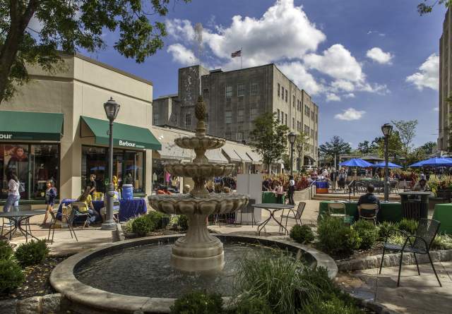 Suburban Square located in Ardmore, PA. The fountain is located in the middle of the shopping district. In the background is the storefront of Barbour. Vendors are seated at tables talking to customers. Many individuals are seen walking around enjoying the warm weather or seated outside for dining at the tables with blue umbrellas.