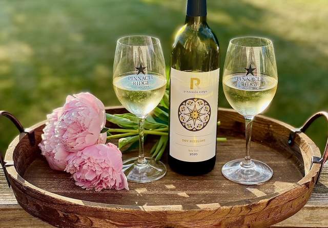 Two glasses of white wine poured into wine glasses featuring the Pinnacle Ridge Winery logo are placed on a serving tray with pink flowers and the white wine bottle.