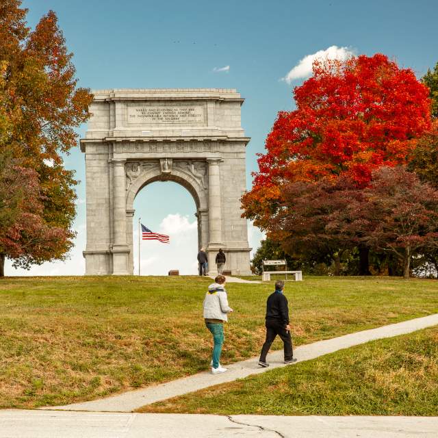 An older male and female walk casually up a hill in front of the monument arch at the Valley Forge National Historic Park surrounded by trees with fall foliage.