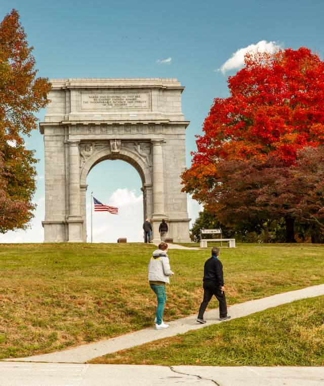 An older male and female walk casually up a hill in front of the monument arch at the Valley Forge National Historic Park surrounded by trees with fall foliage.