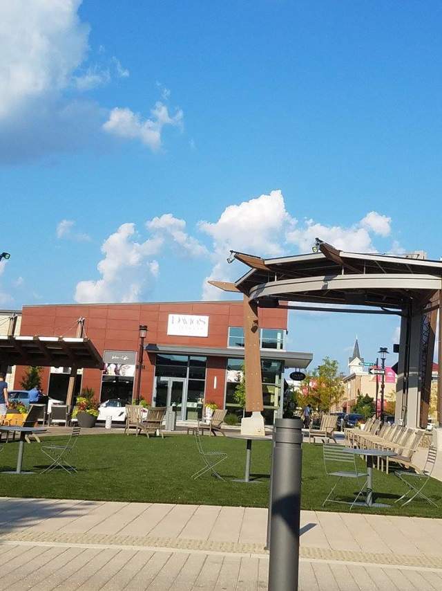 Outdoor plaza with modern buildings, seating areas, and a central pergola under a bright blue sky.