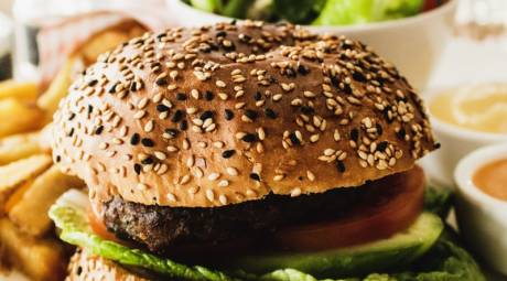 A close-up of a sesame seed bun hamburger with lettuce, tomato, and cucumber, served with french fries and dipping sauces.