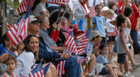 A diverse group of onlookers watch the Skippack Fourth of July Parade from the sidewalk. Numerous spectators are waving American Flags and wearing variations of red, white, and blue.