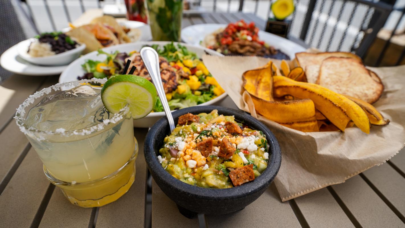A colorful spread featuring guacamole in a stone bowl, plantain chips, a margarita, and various vibrant dishes on a table.
