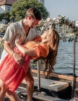 Couple shares joyful moment on dock beside floral-adorned gondola, celebrating with champagne by waterfront