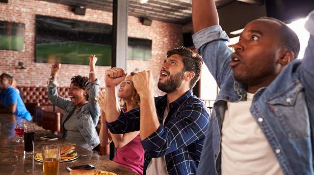 Friends celebrating and cheering at a sports bar while watching a game, with burgers, fries, and drinks on the table.