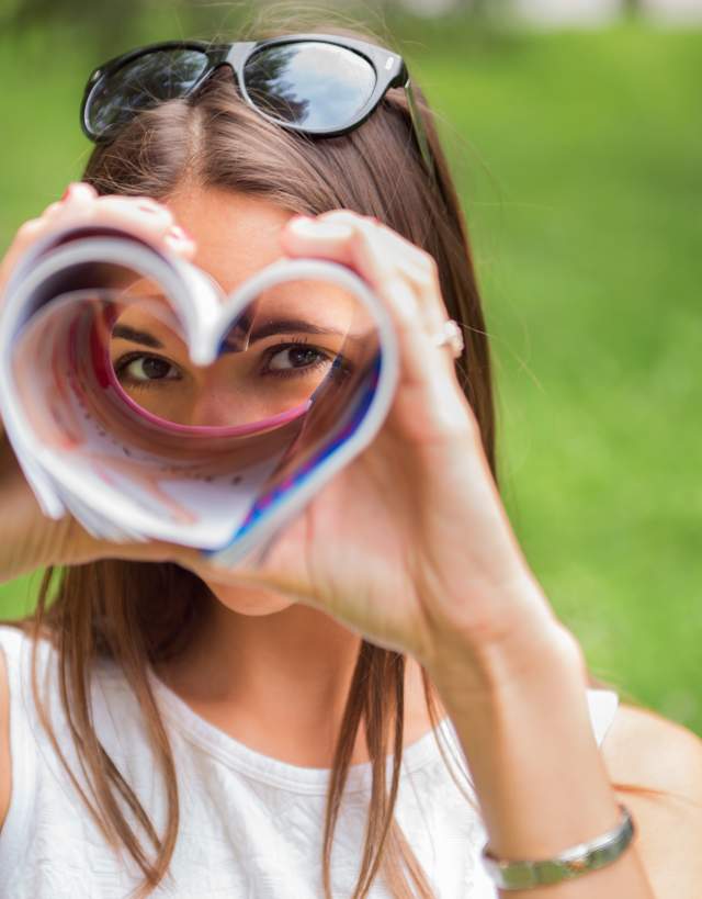 Woman with sunglasses on her head, looking through a magazine folded in the shape of a heart