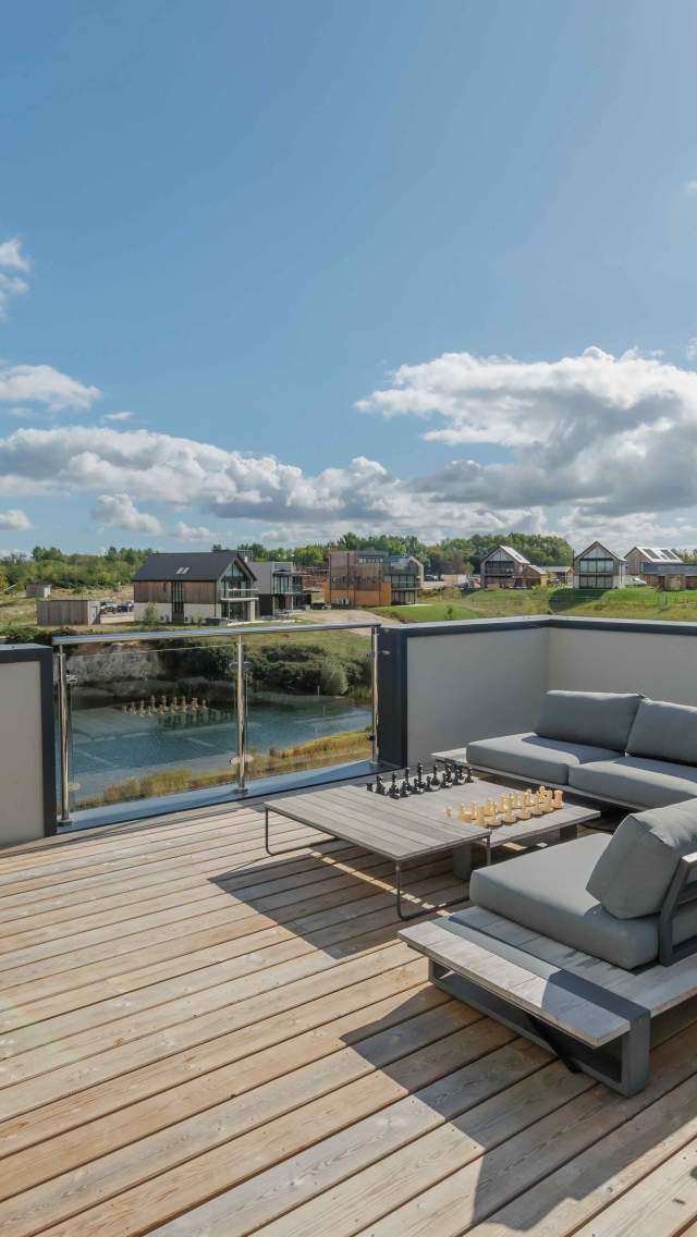 A modern rooftop deck with grey lounge furniture and wooden chairs overlooks a coastal landscape of houses and greenery under a bright, cloudy sky.