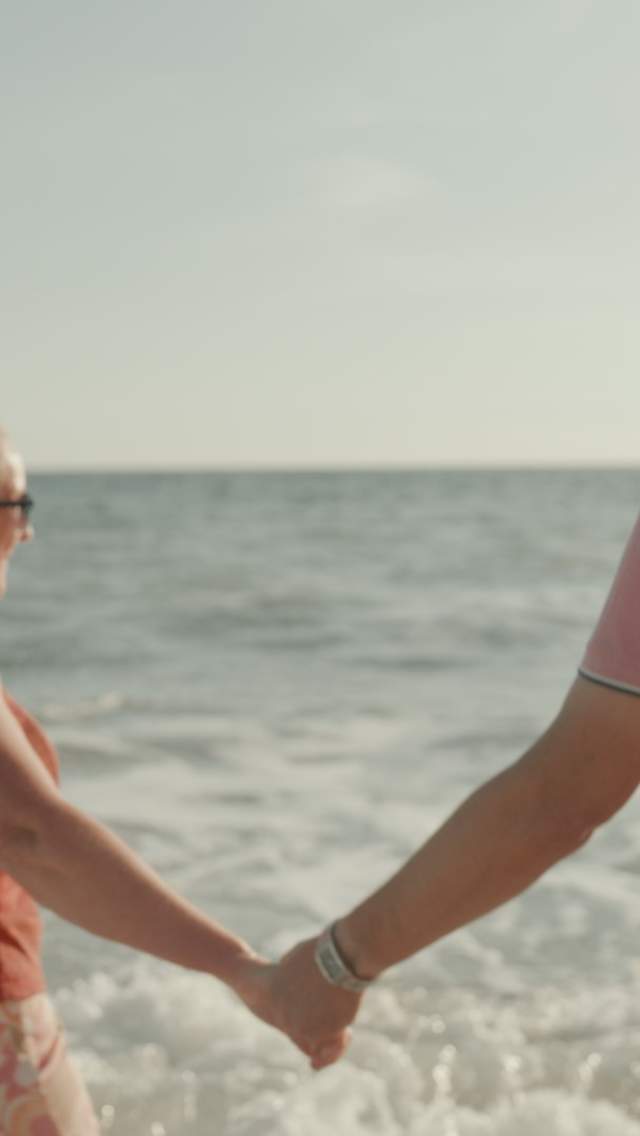 Man and woman holding hands as they stand in the waves on a beach in Dorset