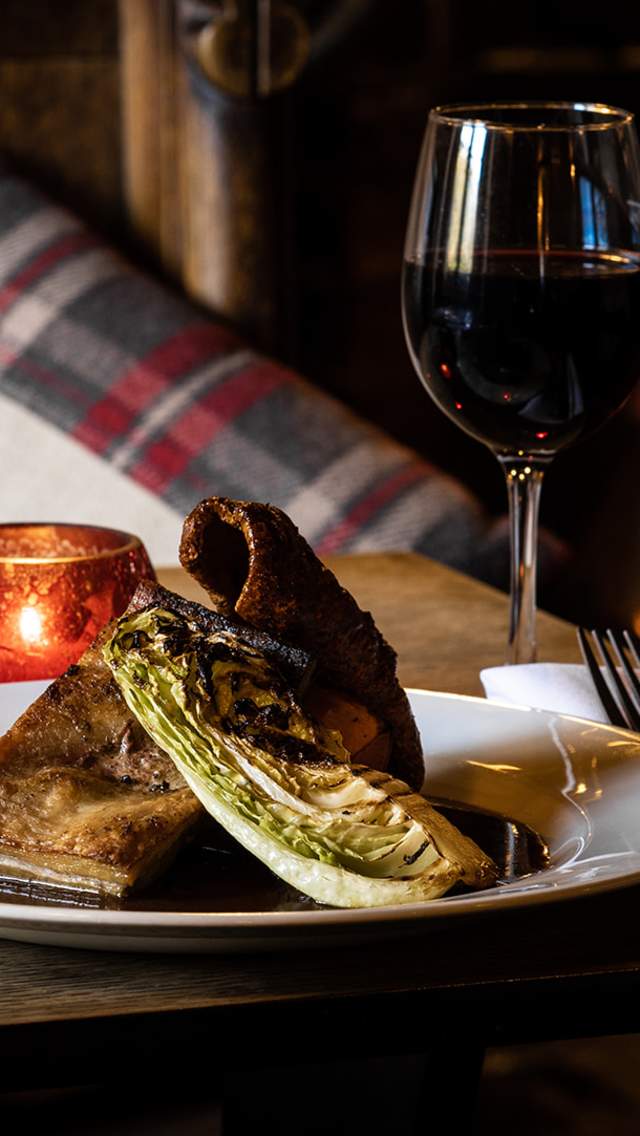 A plate of food and a glass of red wine on a table in front of a log fire at the Brace of Pheasants pub in Dorset