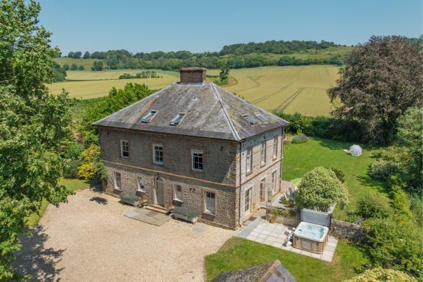 A large stone house with a gravel driveway in the foreground sits amidst lush green fields and trees under a clear blue sky in the background.