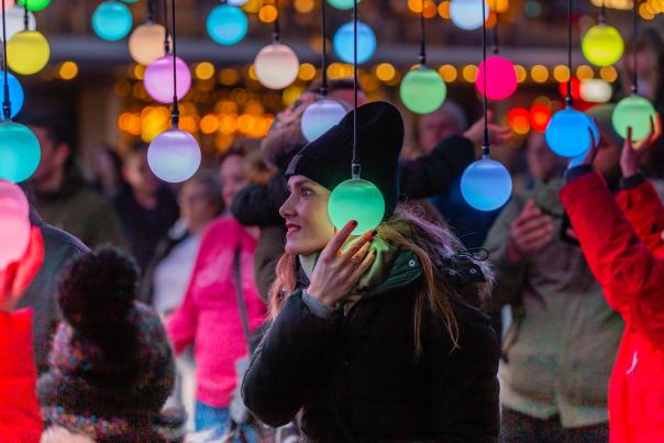 A woman in a black hat and jacket stands in the foreground, surrounded by glowing colorful spheres and a blurred crowd in the background.