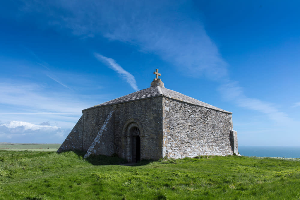 A stone chapel with an arched doorway stands on a grassy hill under a bright blue sky with wispy clouds, overlooking the sea in the distance.