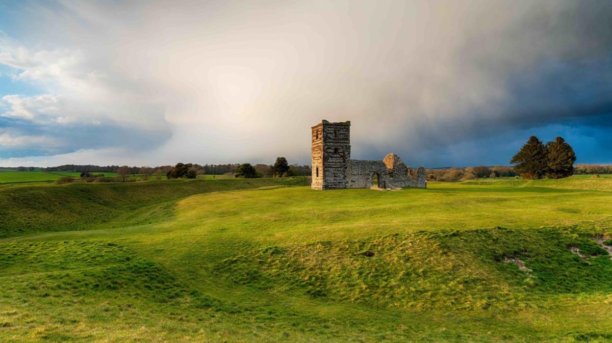 Rolling green hills with a ruined stone tower standing under a dramatic, cloudy sky.