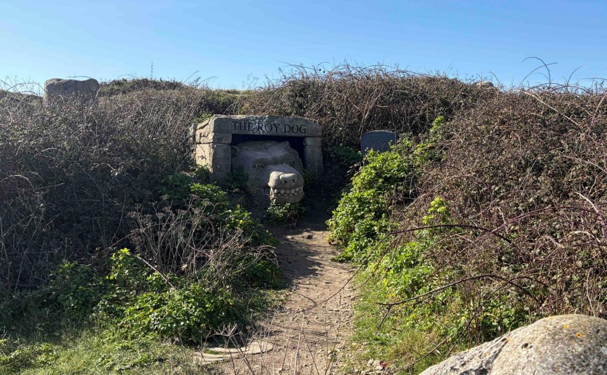 A dirt path leads through overgrown bushes to a stone structure with "THEY'S DOG" carved above its entrance, under a clear blue sky.
