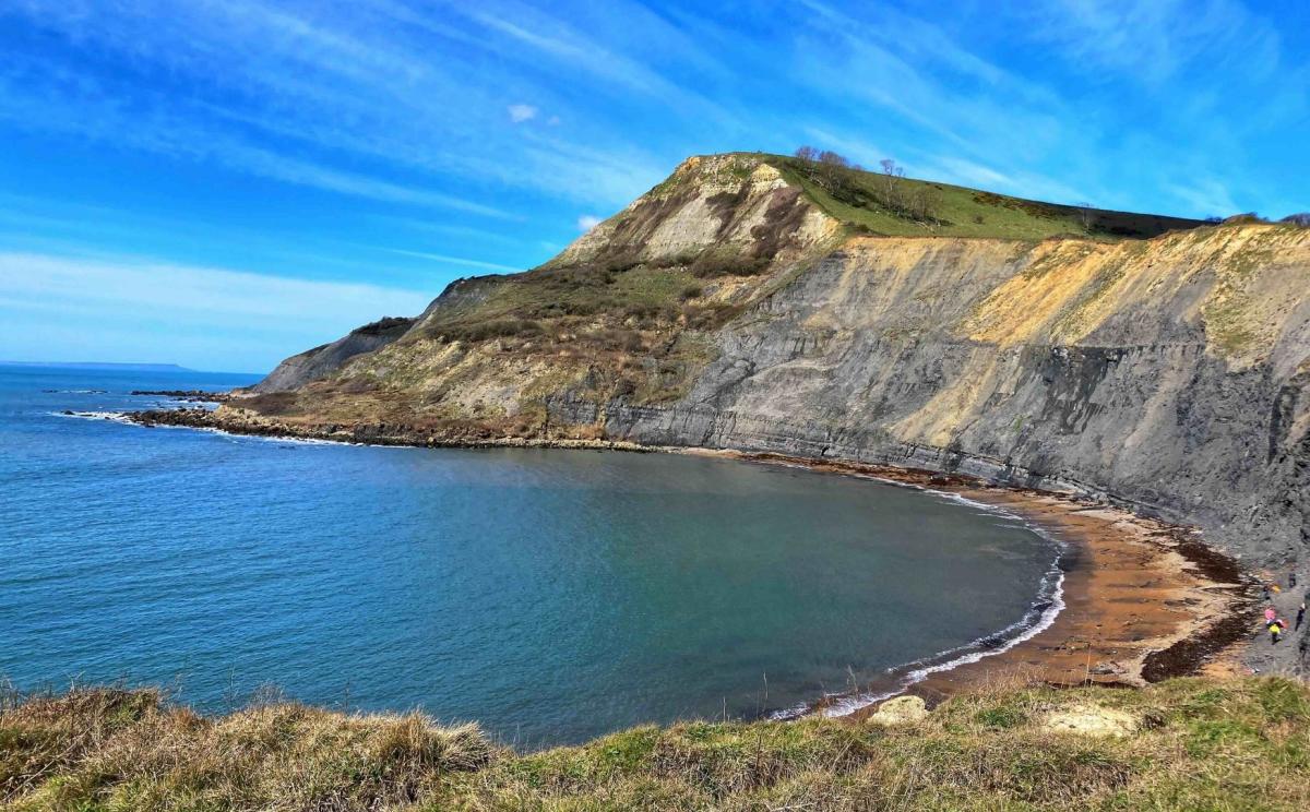 A calm, blue bay with a sandy beach curves around the base of a dramatic, layered cliff face under a bright blue sky with wispy clouds.