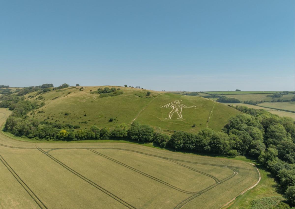 A giant white figure is etched into a grassy hillside in the background, with cultivated fields in the foreground.