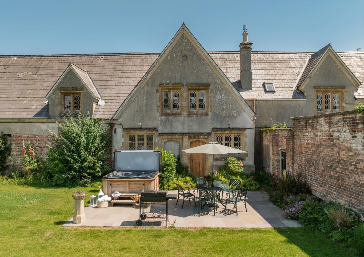 A patio with a hot tub, grill, and outdoor dining set sits in the foreground before a large, stone country house with a brick wall to the right.