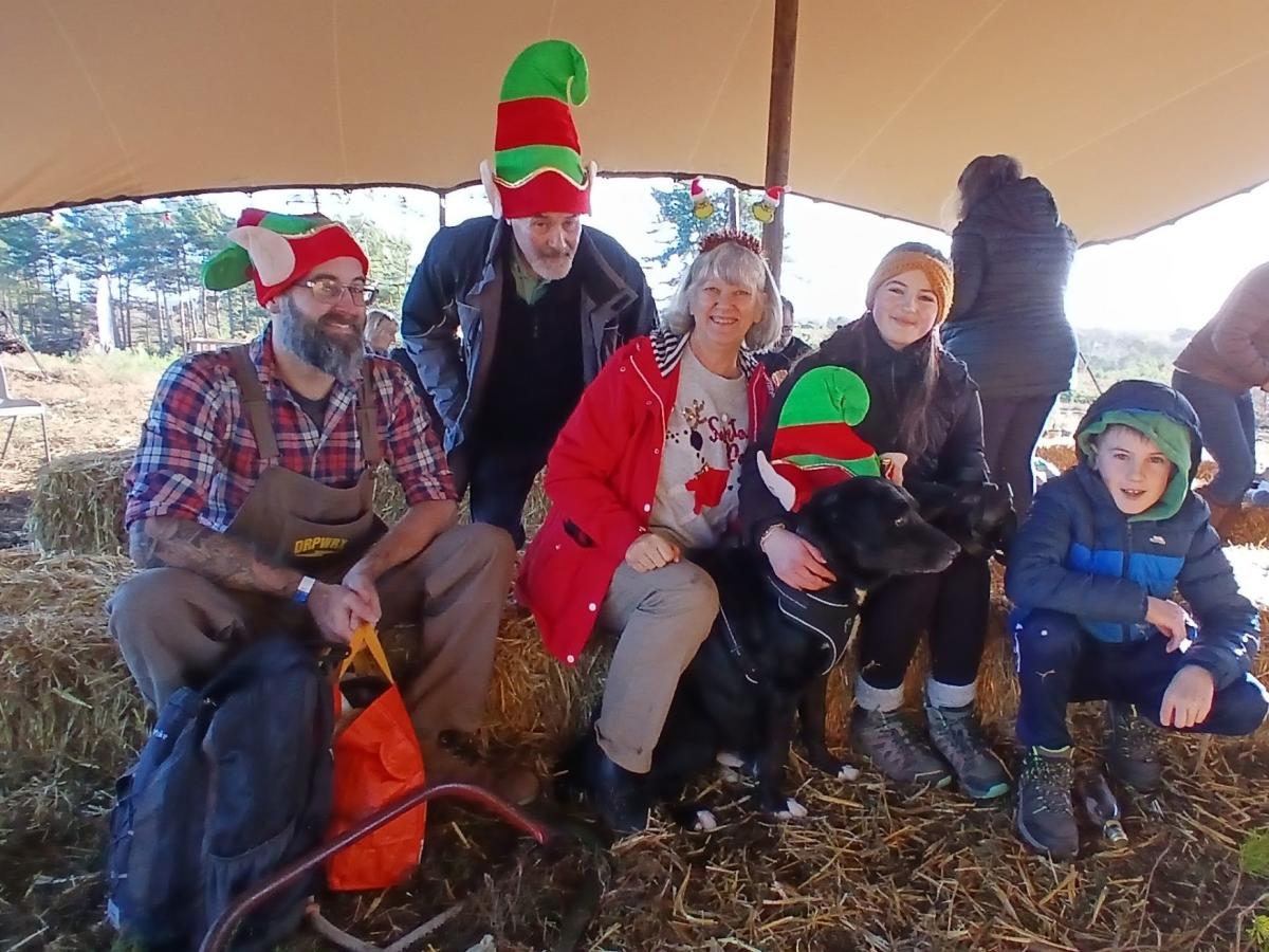 A group of people and a dog wearing festive hats sit on hay bales under a tan canopy.